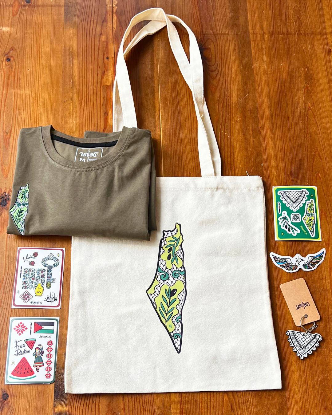 Uniart Palestine Package - T-shirt, tote bag, 3 sticker sheets, unique sticker, and keychain with hand-drawn Palestinian designs by Hala Jafar.
باقة فلسطين من يوني آرت - تيشيرت، حقيبة قماشية، 3 أوراق ستيكرات، ستيكر مميز، وميدالية مفاتيح مع تصاميم فلسطينية مرسومة يدوياً من حلا جعفر."